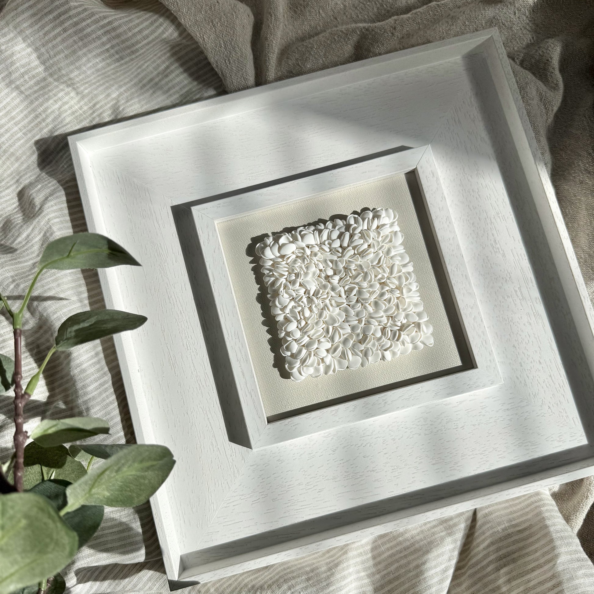 Decorative square piece with textured design in a white frame on a textured surface with green leaves.